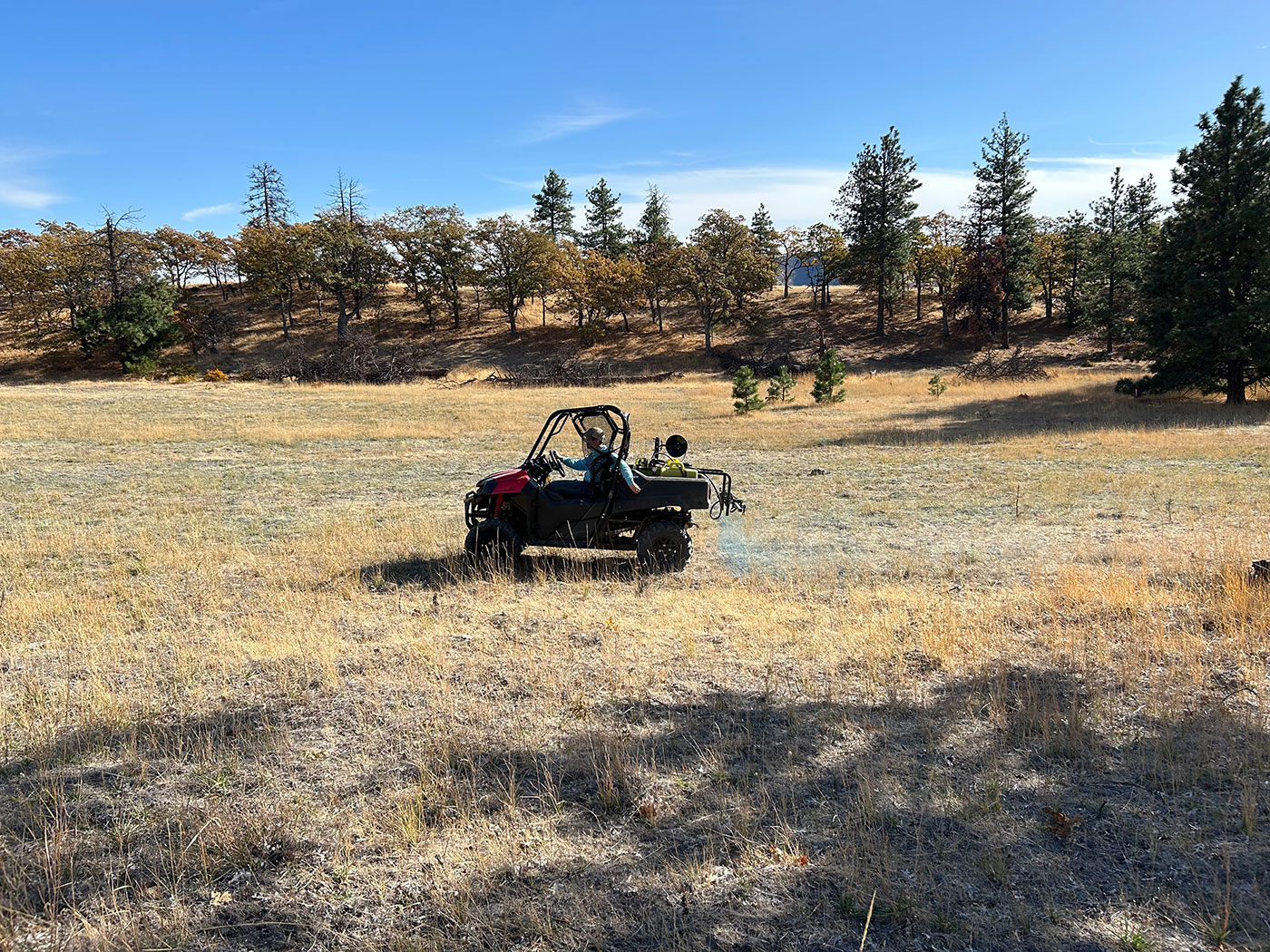 A small off-road utility vehicle is parked in a dry, grassy field surrounded by scattered trees under a clear blue sky. Various bags and gear are attached to the vehicle.