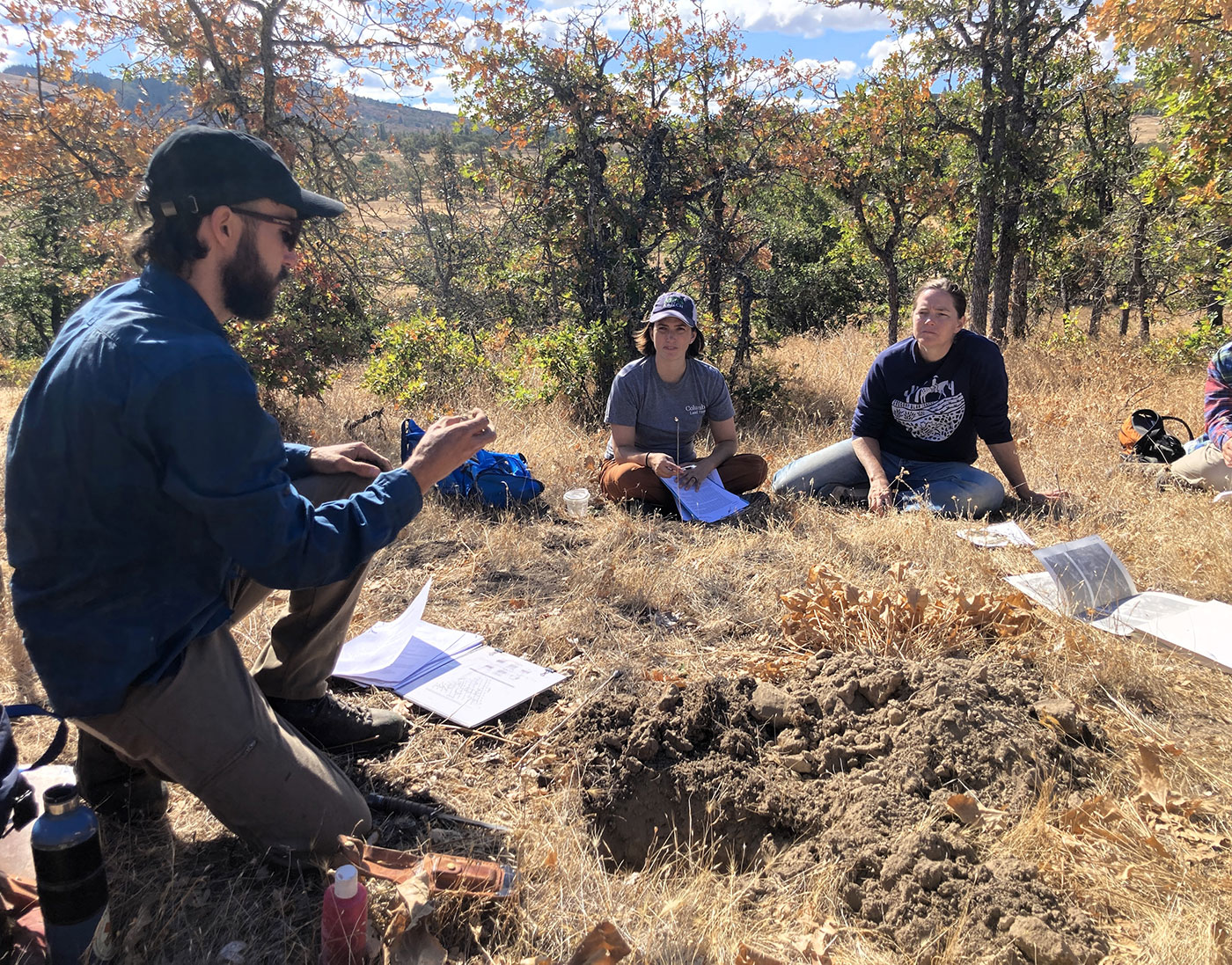 Three people sit on dry grass in a sunny, wooded area, surrounded by papers and notebooks. One person gestures while talking, while two others listen and take notes. Exposed soil and scattered belongings are visible nearby.