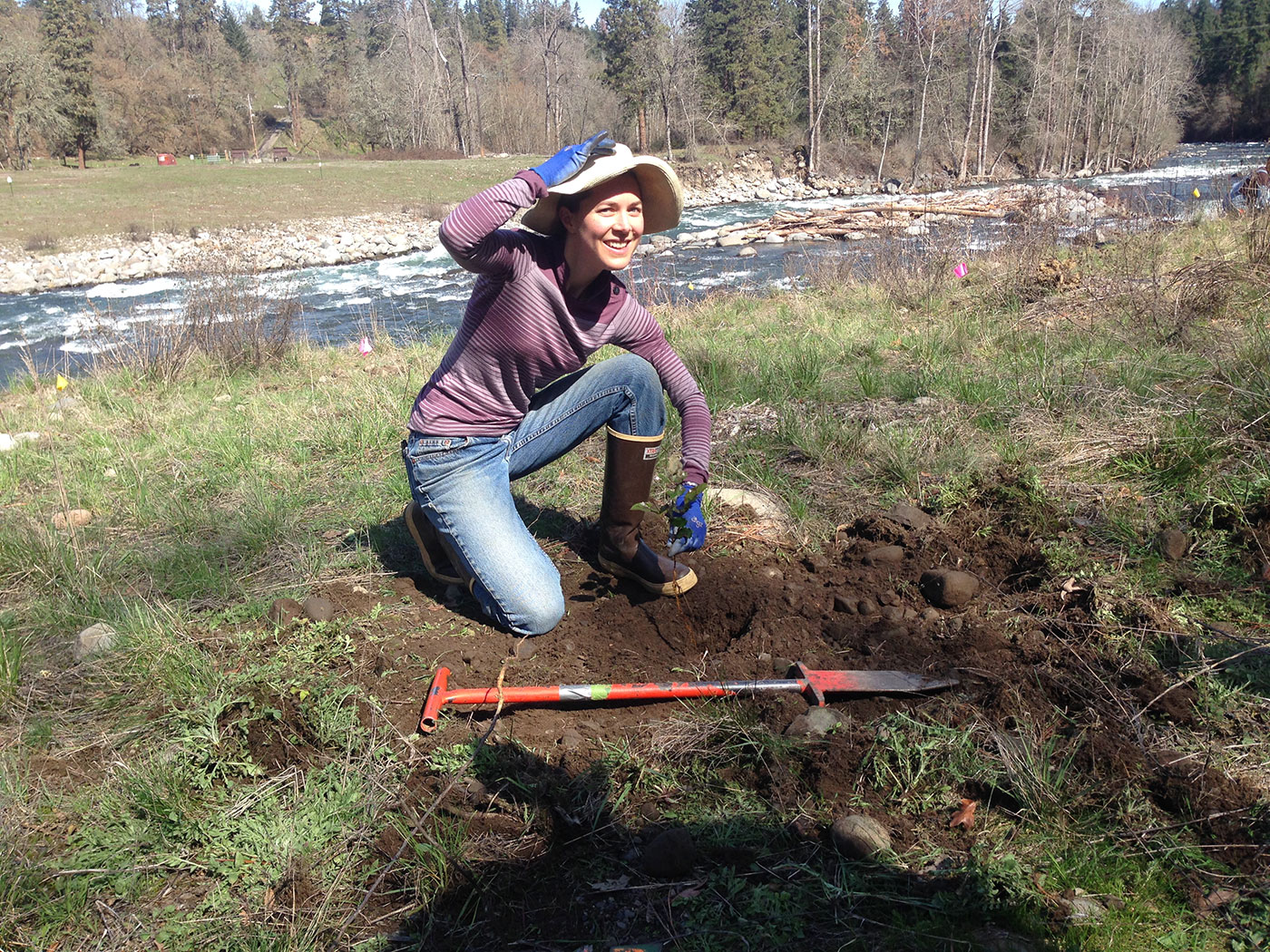 A person kneels on grass near a river, smiling and holding their wide-brimmed hat. They wear boots and gloves, with a long-handled tool lying on the ground in front of them. Trees and a flowing river are in the background.