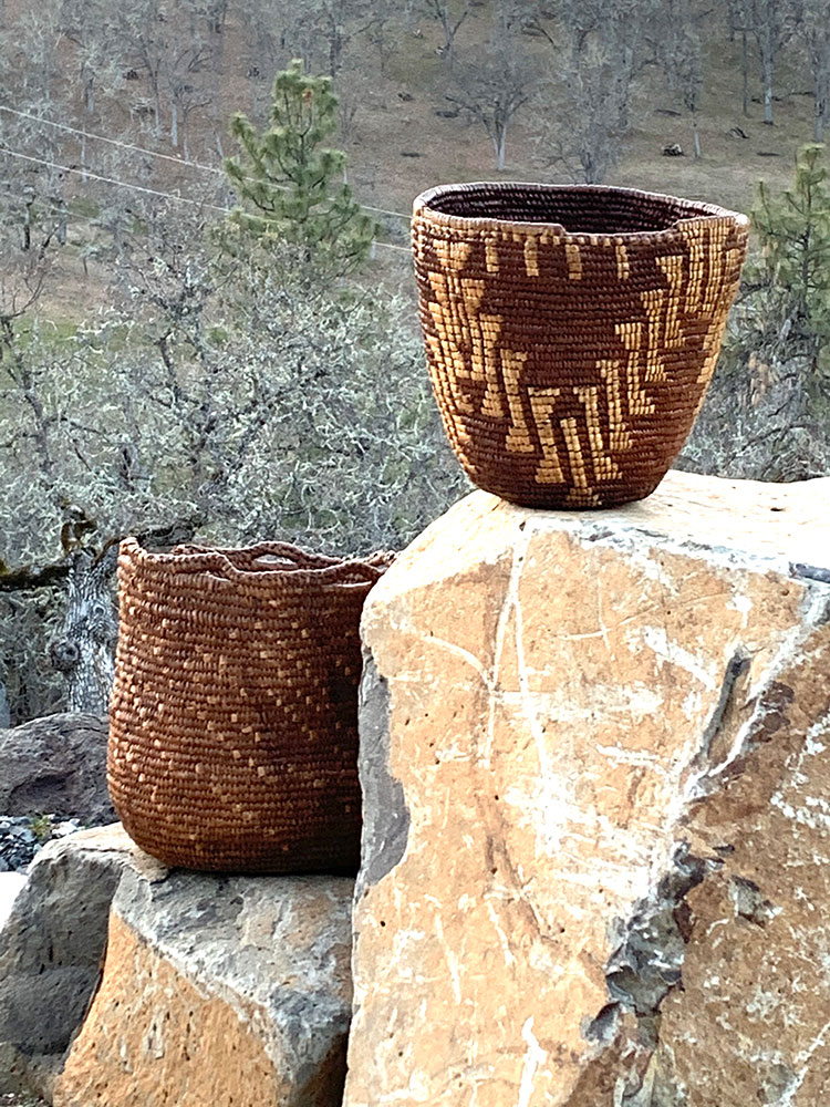 Two woven baskets sit outdoors on large tan and brown rocks, with a background of leafless trees and dry grass. One basket features a zigzag pattern and the other has a simpler, uniform design.
