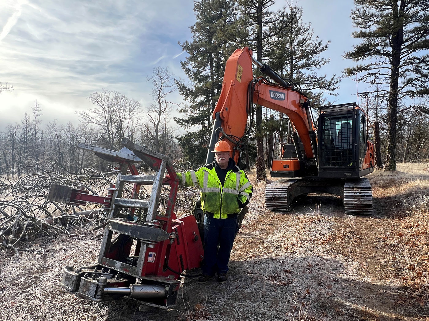A person in a yellow safety jacket and orange hard hat stands beside logging equipment and an orange excavator in a frosty forest clearing, with fallen branches and tall trees in the background.