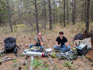 Two researchers sitting on the ground in an oak woodland use machines to test oak leaf samples.